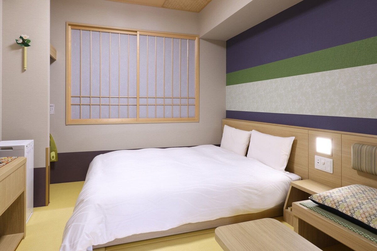 A hotel room with a bed, bedside table, TV, and a window with vertical blinds.