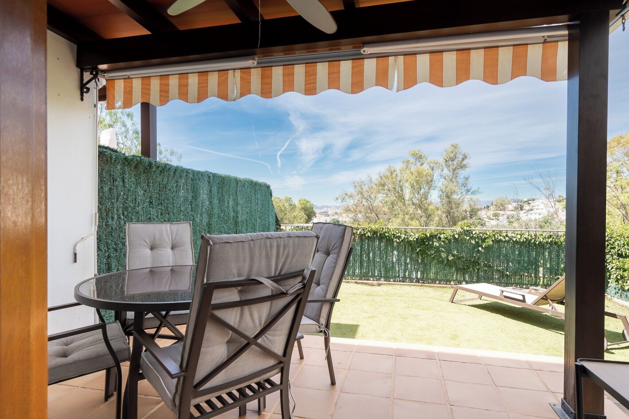 Une terrasse extérieure avec une table ronde en verre et des chaises rembourrées, un auvent à rayures et une vue sur une pelouse et des haies bien entretenues.
