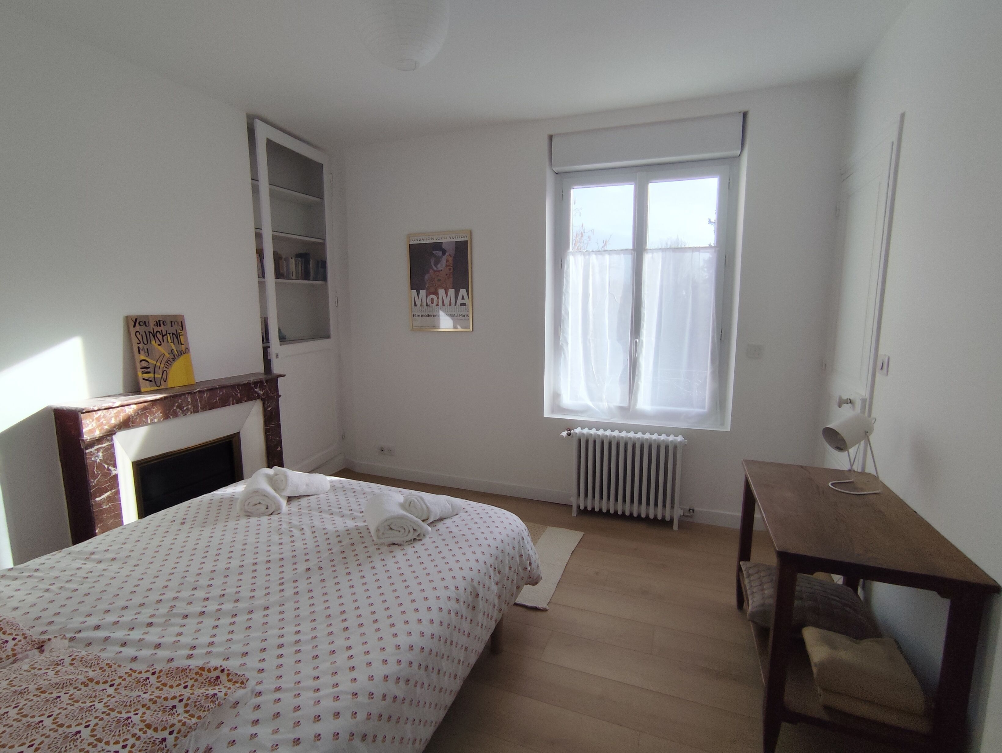 A bedroom with a bed, a radiator, a window with curtains, a small table, and a shelf with books.
