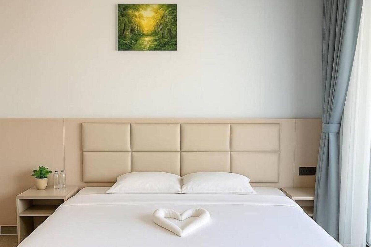 A neatly made bed with a heart-shaped pillow, a bedside table with a plant and bottles, and a painting on the wall.