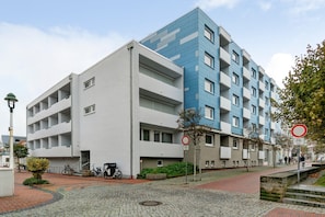 Un immeuble d’appartements moderne aux façades aux couleurs variées, allant du blanc au bleu, situé sur une rue pavée, avec un vélo garé devant.
