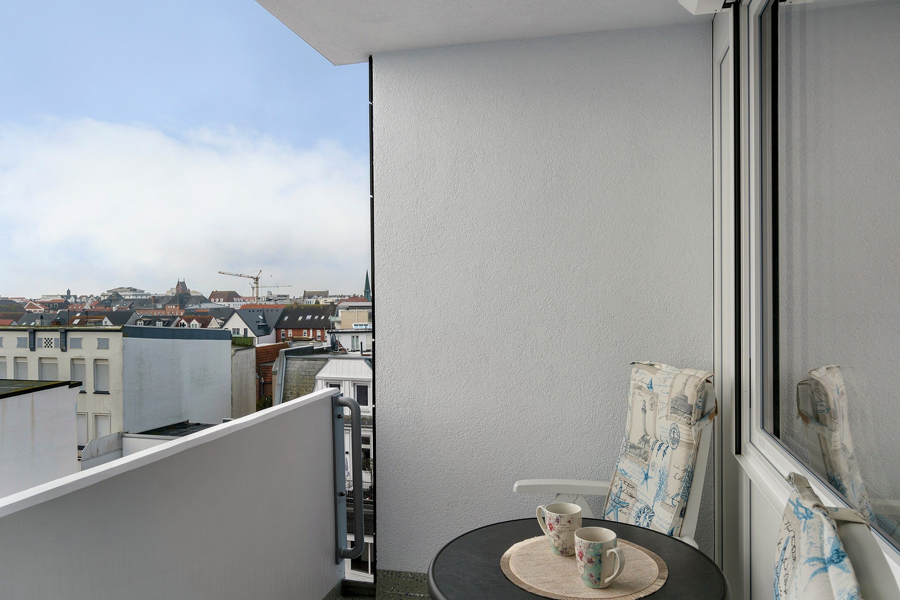 A balcony with a table and two cups, overlooking a cityscape with buildings and a crane.