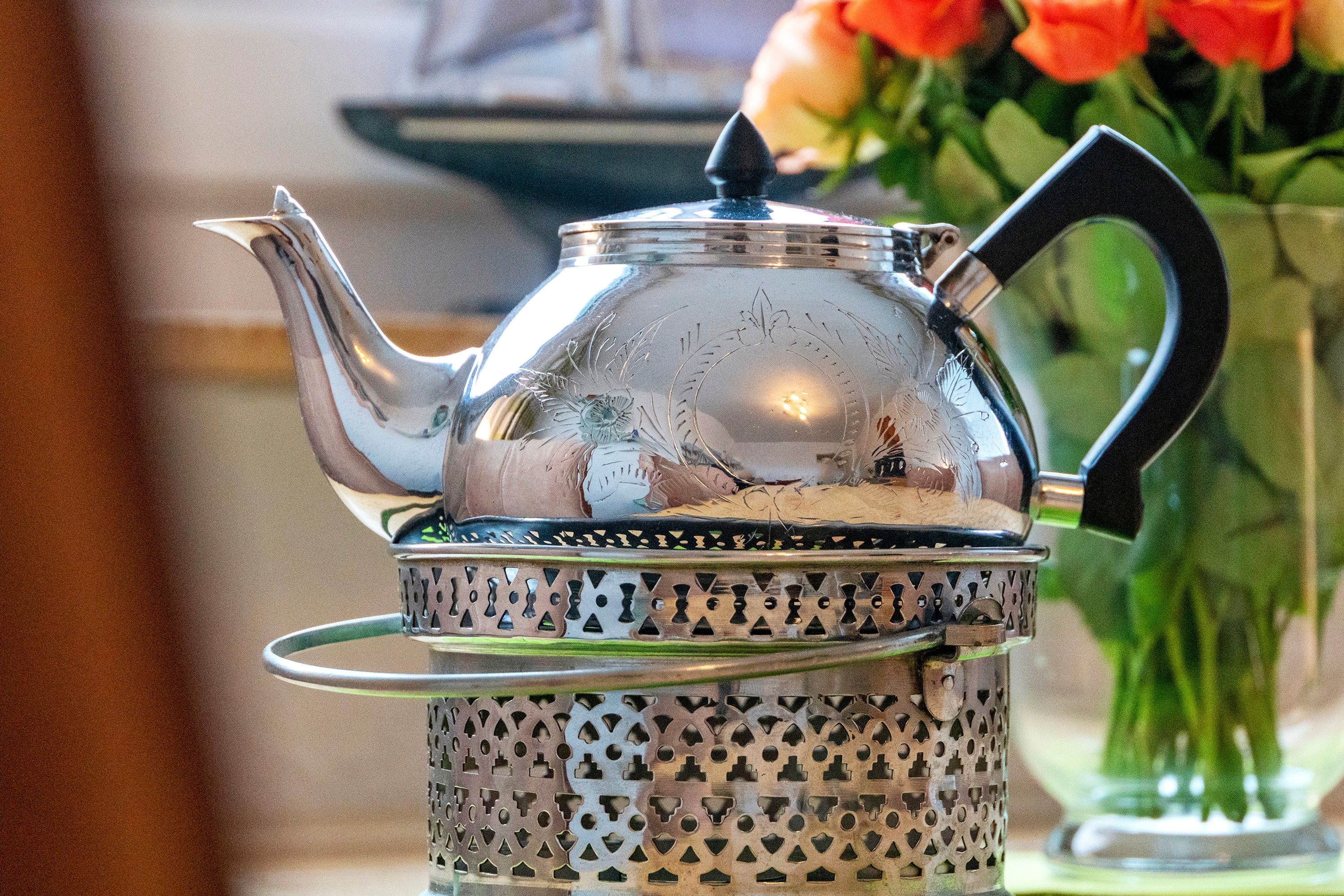 A silver teapot with a handle and spout, placed on a metal strainer with a patterned design.