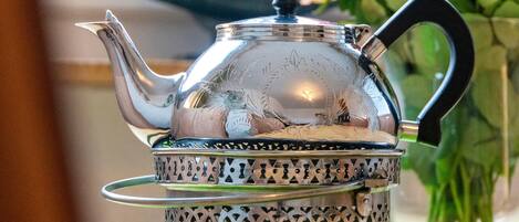 A silver teapot with a handle and spout, placed on a metal strainer with a patterned design.
