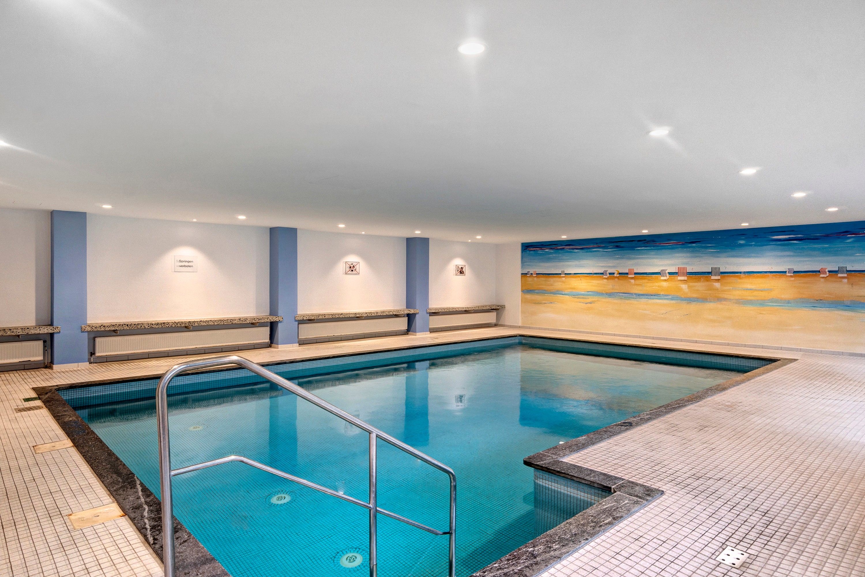 Une piscine intérieure à l’eau d’un bleu limpide, avec un escalier en métal et un sol carrelé.