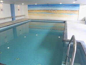 Piscina interior com vista para um mural de praia, piso de azulejos e guarda-corpo de metal.
