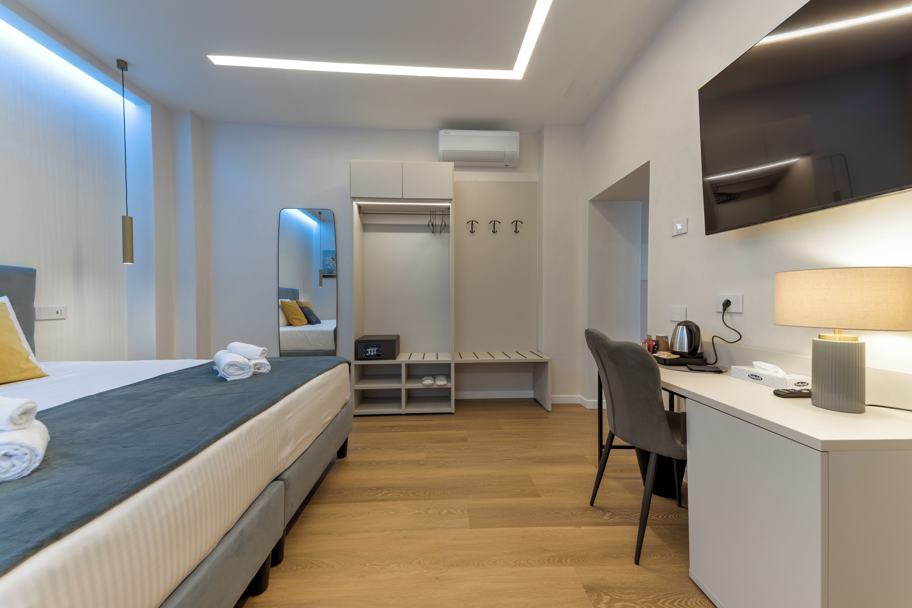 A modern hotel room with a bed, desk, chair, wardrobe, and a TV mounted on the wall.