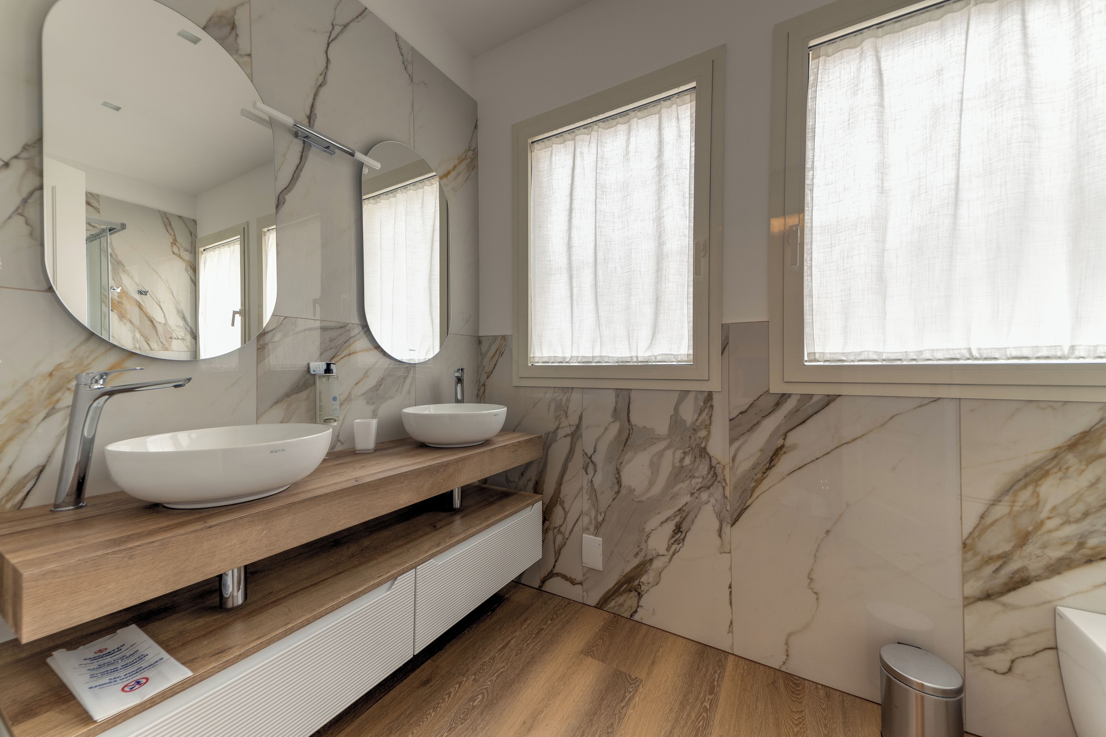 A modern bathroom with a marble wall, a large mirror, two sinks, and a window.