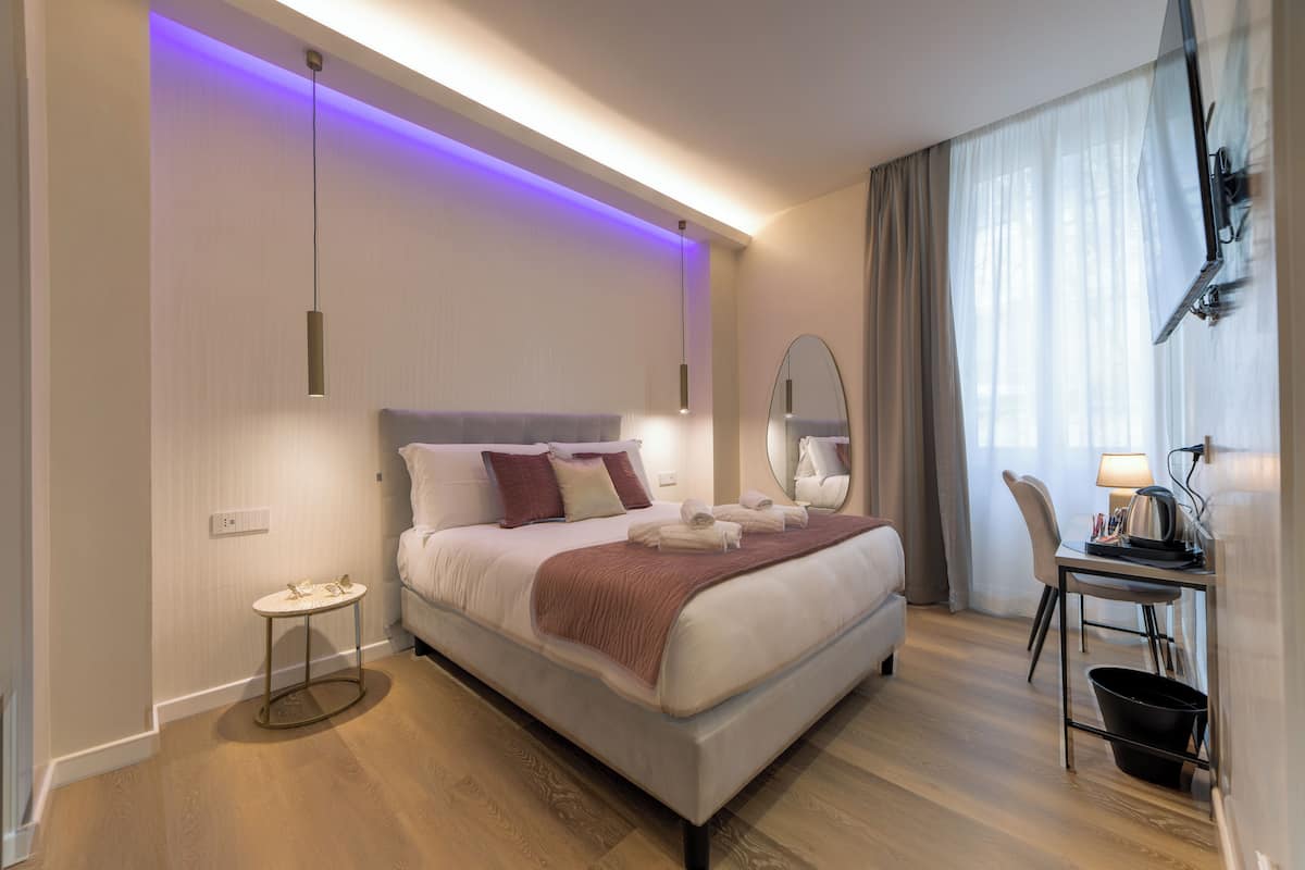 A modern hotel room with a bed, bedside table, mirror, and a desk with a chair.