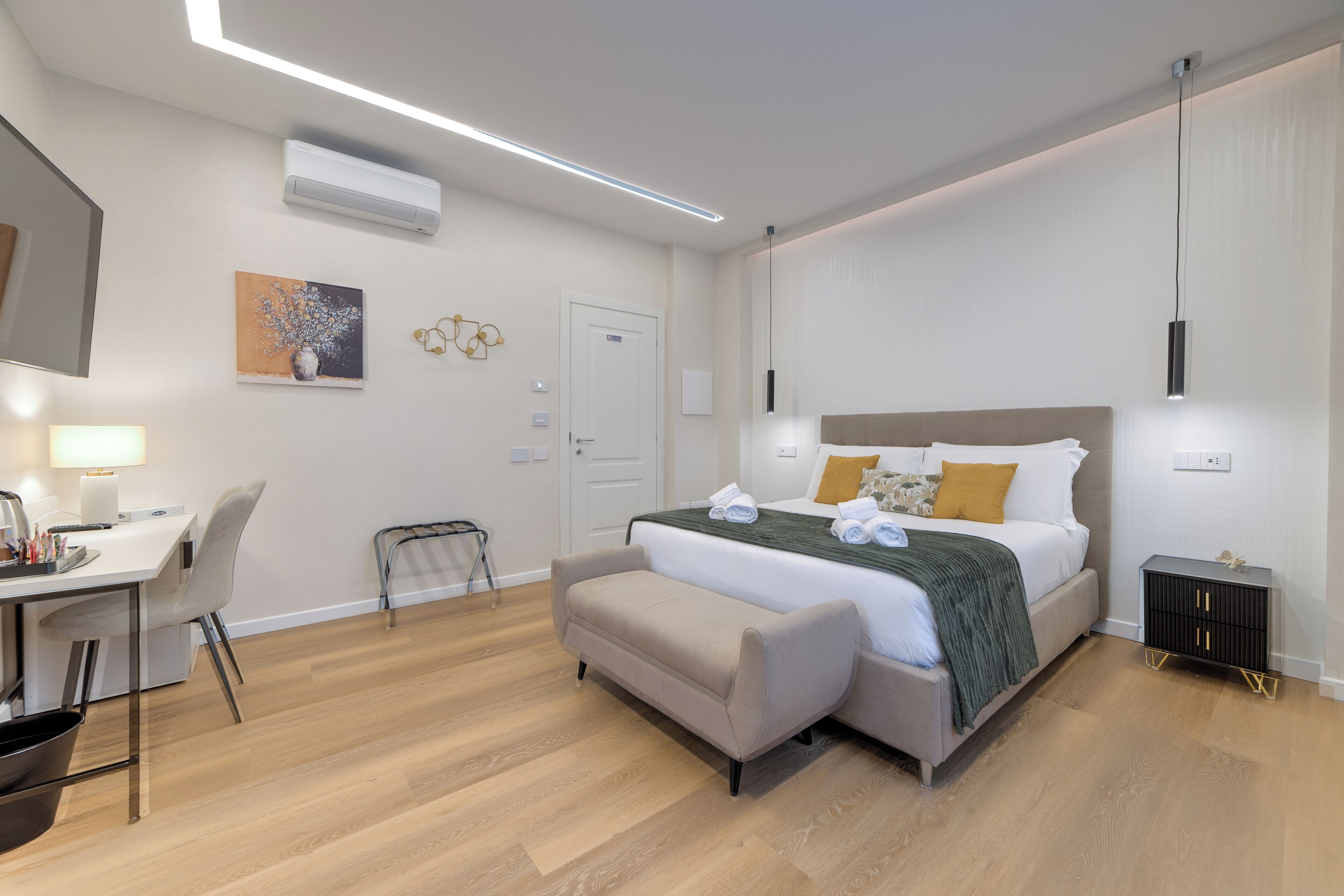 A modern hotel room with a bed, bedside table, desk, chair, and an air conditioning unit.