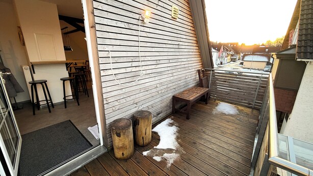 Une terrasse en bois avec un banc et des tabourets, une porte donnant sur un espace intérieur et une vue sur l’extérieur.