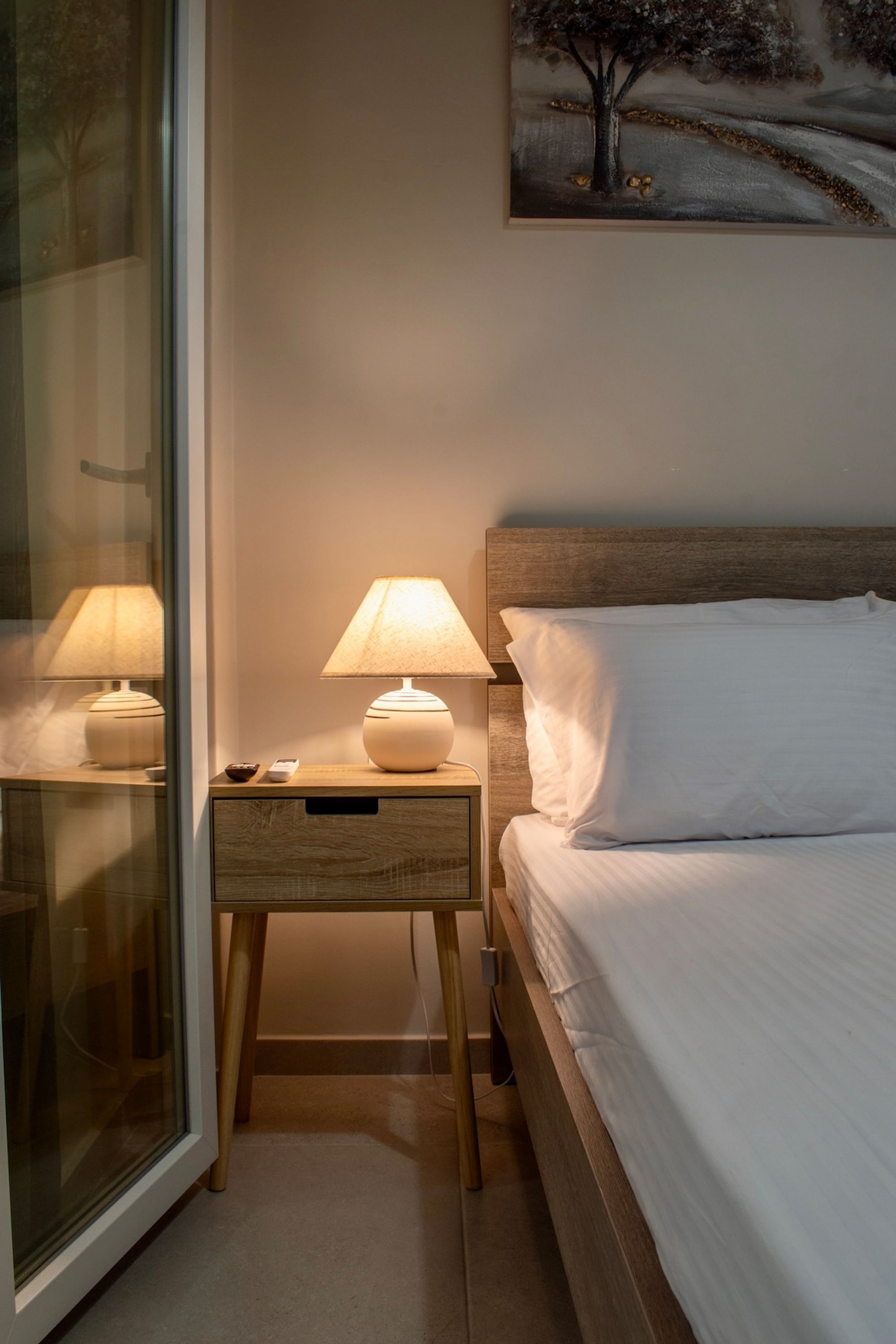 A hotel room with a bed, bedside lamps, a nightstand, a painting on the wall, and a glass door.