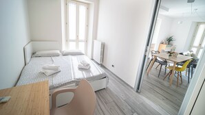 Een slaapkamer met een bed, nachtkastje, stoel, raam en uitzicht op de eetruimte.