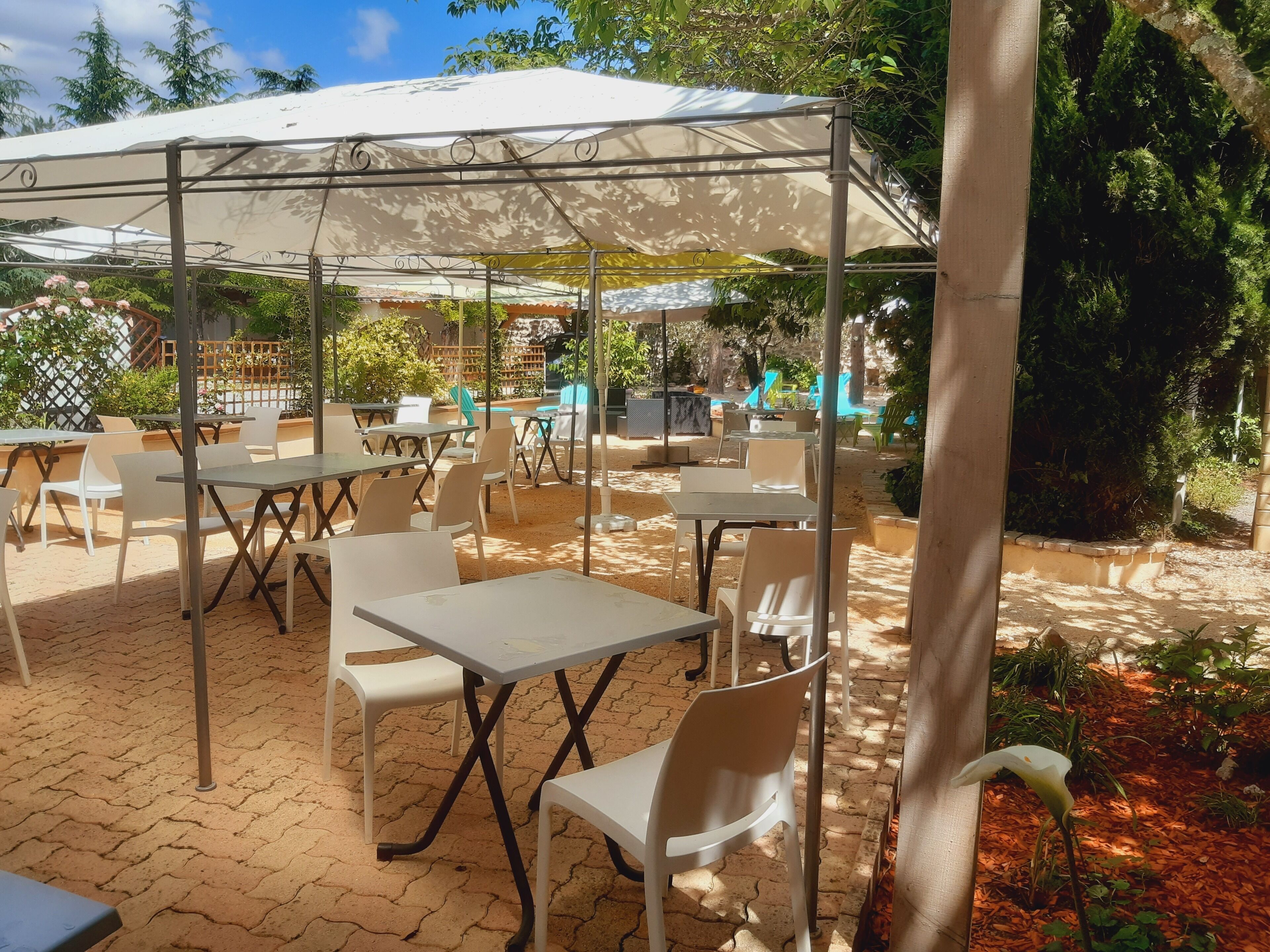 An outdoor patio with tables and chairs, covered by a canopy, surrounded by greenery.