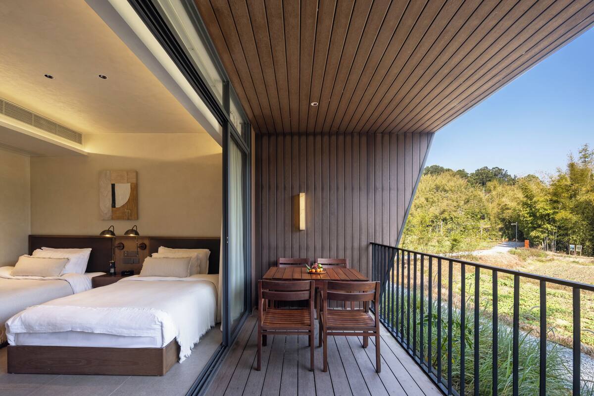 A balcony with a wooden table and chairs, a bed with white bedding, and a view of the outdoors.