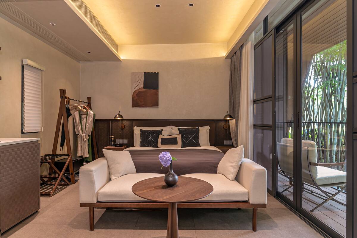 A bedroom with a bed, bedside tables, a sofa, a small table with a vase, and a view of a bamboo garden through large windows.