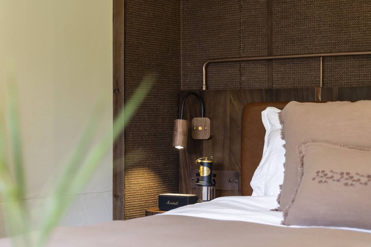A neatly made bed with a headboard, a small bedside table with a lamp and a device, and a textured wall.