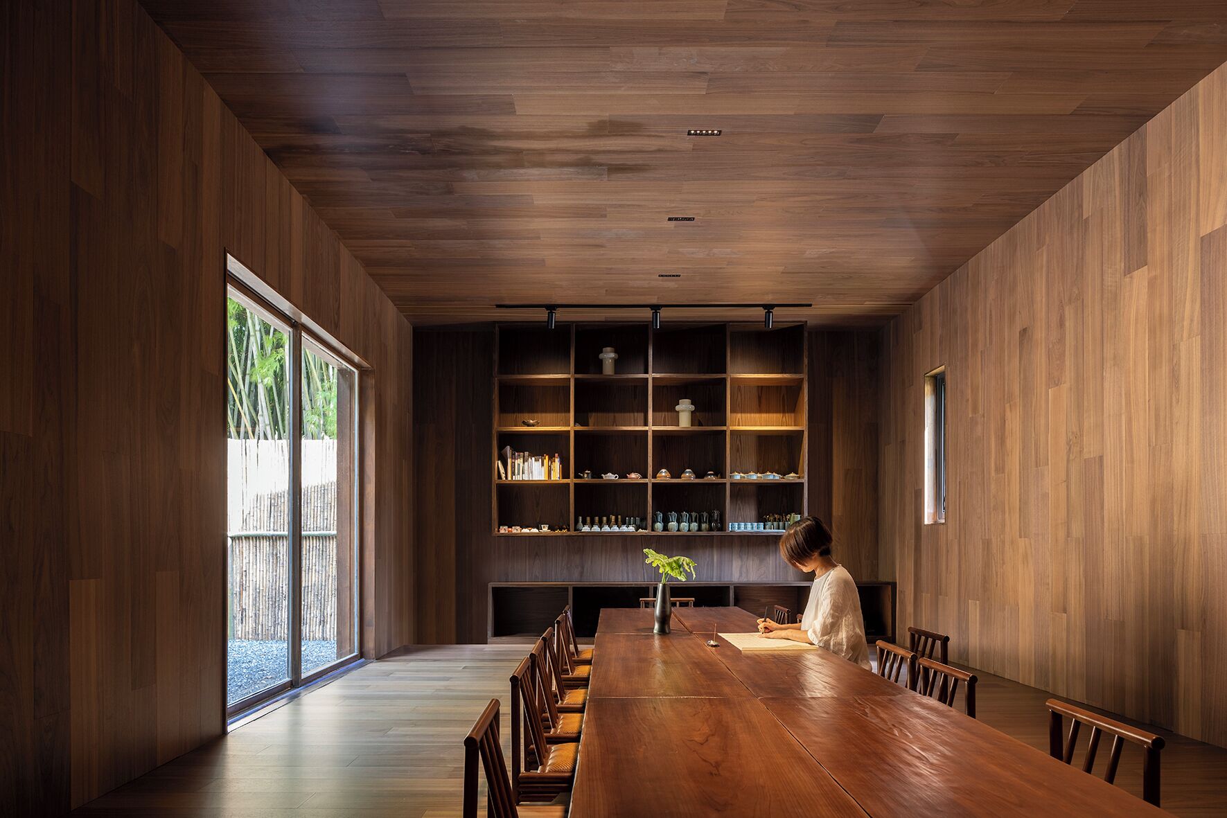 Une salle à manger moderne avec une longue table en bois, des chaises et une bibliothèque remplie de livres et d’objets décoratifs.