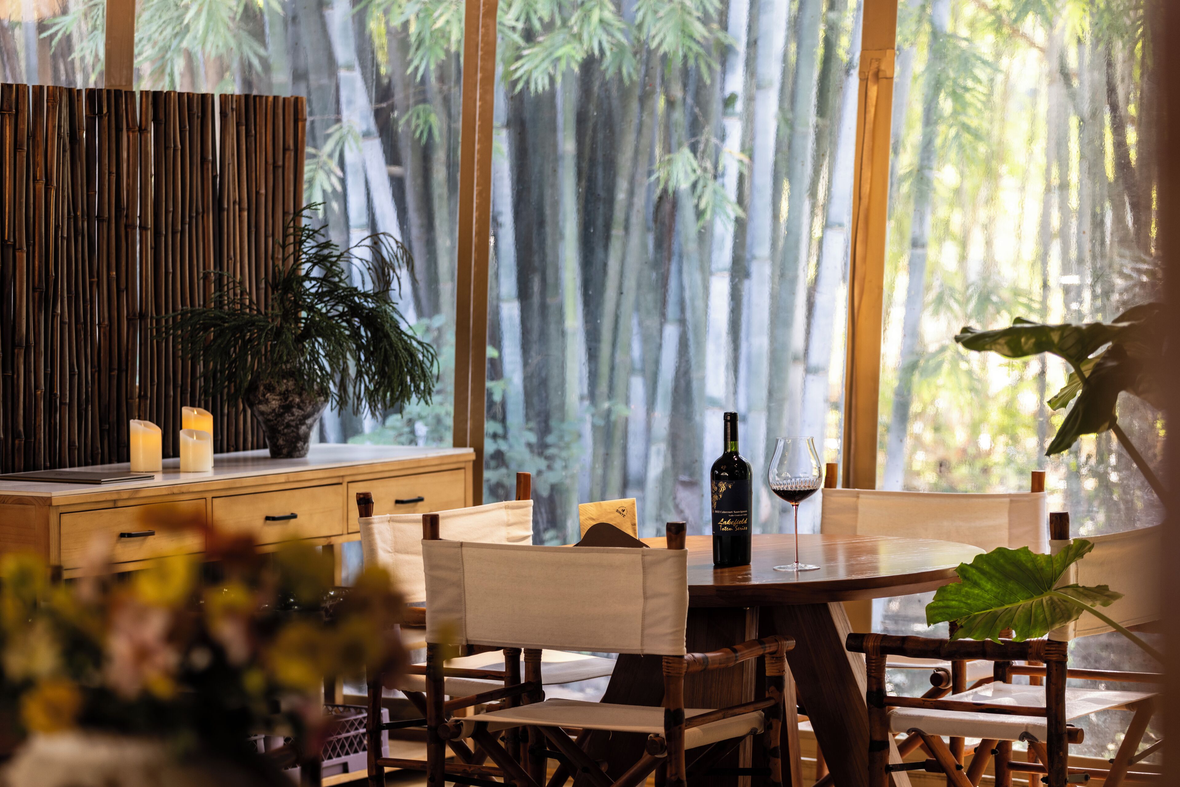 A dining area with a round table, chairs, candles, a bottle of wine, and a large window with a bamboo forest view.