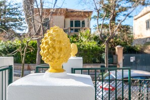 A white pedestal with a yellow decorative object, a green metal fence, and a building with a tiled roof in the background. - Mondello sea life garden (PALERMO)
