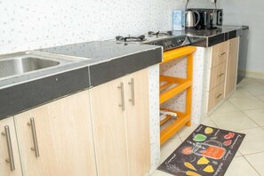 A kitchen with a black countertop, stainless steel sink, wooden cabinets, a yellow shelf, and a floor mat with fruit illustrations.