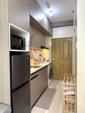A compact kitchen with a built-in microwave, a stainless steel refrigerator, a washing machine, and a wooden door.
