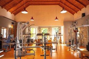 Une salle de sport équipée de divers appareils d’exercice, avec des poutres en bois au plafond et de grandes fenêtres laissant entrer la lumière naturelle.