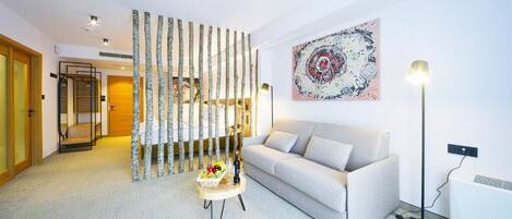 Een moderne woonkamer met een bank, een decoratief wandkunstwerk, een vloerlamp en een klein tafeltje met een fles en fruit.
