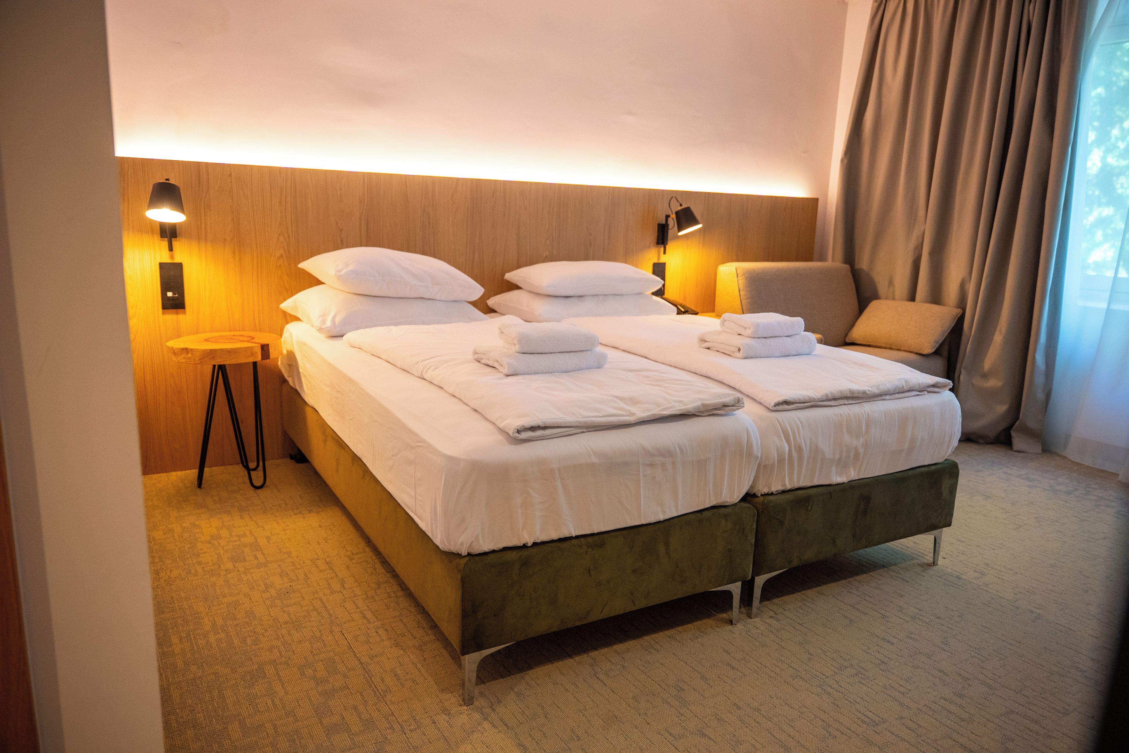 A double bed with white linens and pillows, a bedside table with a lamp, a chair, and a view of the outdoors through a window with curtains.