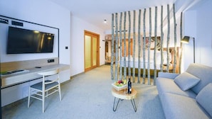 Una moderna camera d'albergo con TV a schermo piatto, una scrivania, un divano, un pannello decorativo a forma di albero e un piccolo tavolino con una bottiglia e della frutta.