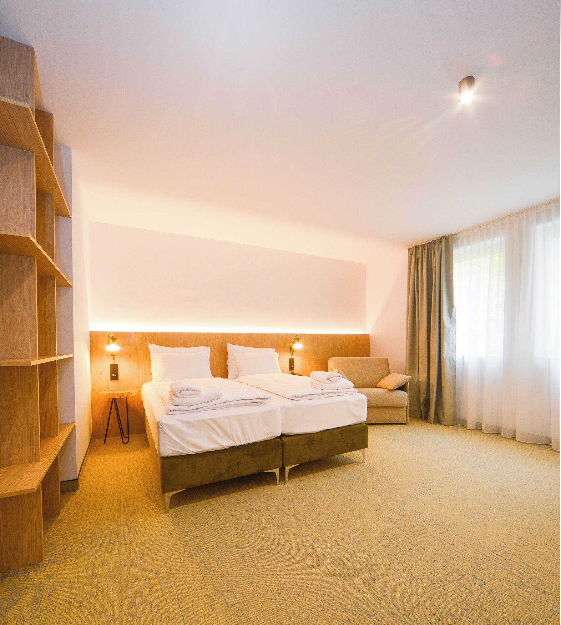 A hotel room with a large bed, a sofa, a wooden bookshelf, and a wall-mounted light.