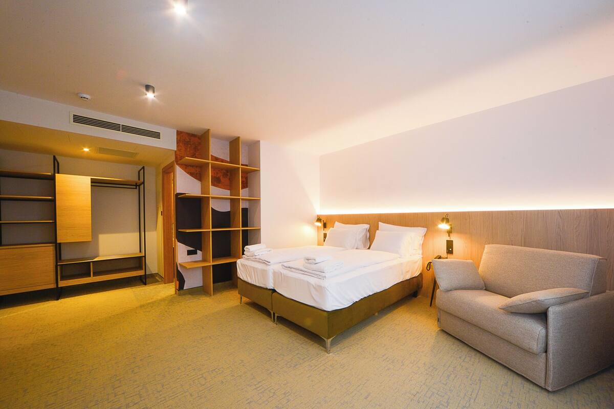 A hotel room with a bed, a sofa, a wardrobe, and a wall-mounted shelf.