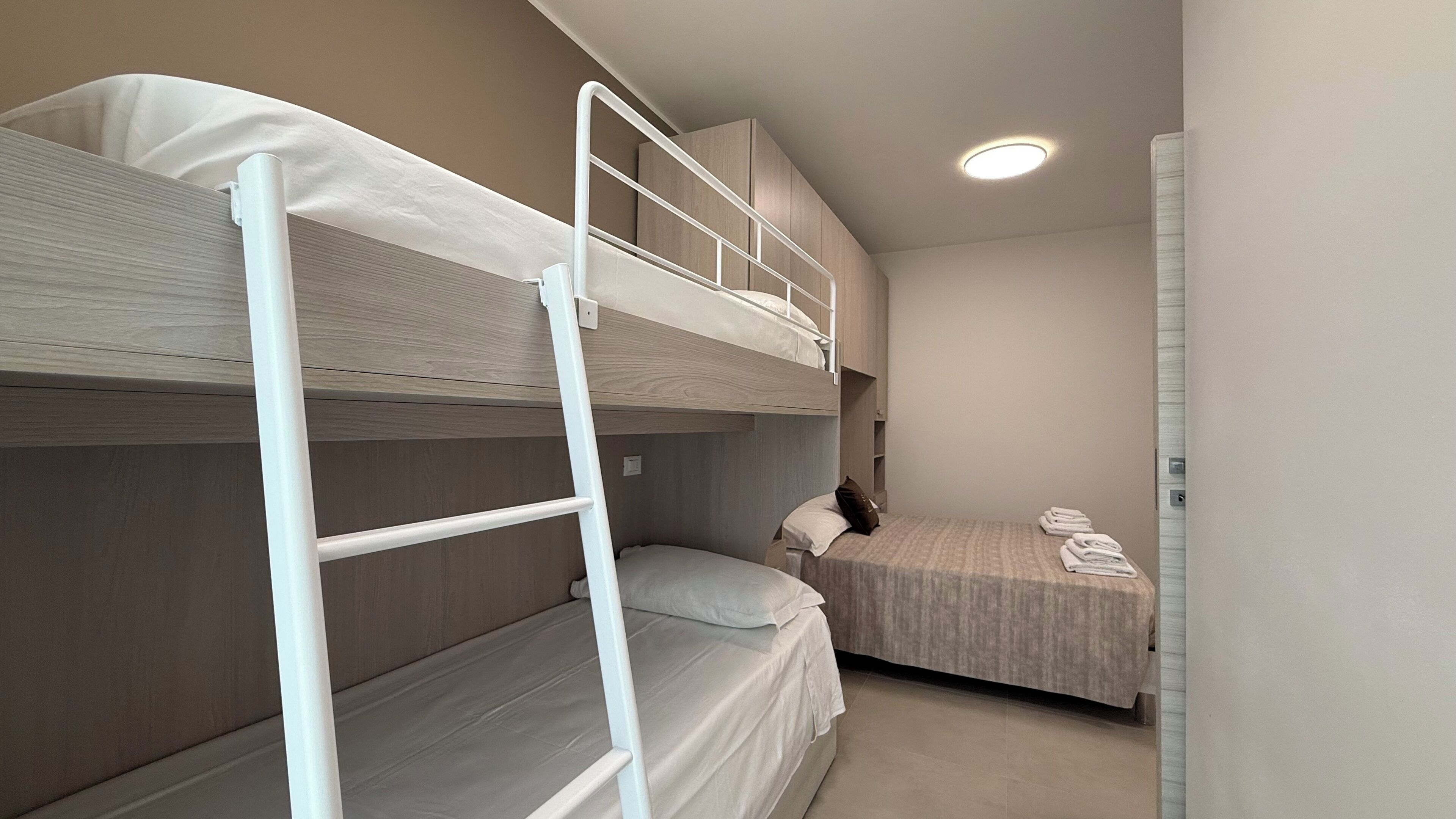 A bunk bed room with a ladder, a lower bunk, and a single bed with a pillow and folded towels.