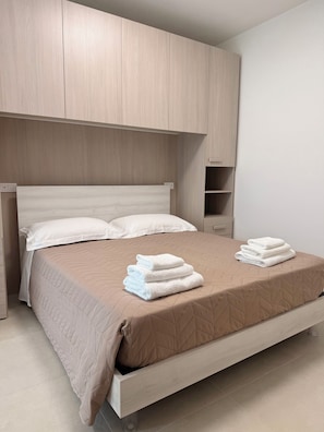 Una cama con una colcha beige acolchada, dos almohadas blancas y una pila de toallas blancas dobladas sobre la cama.