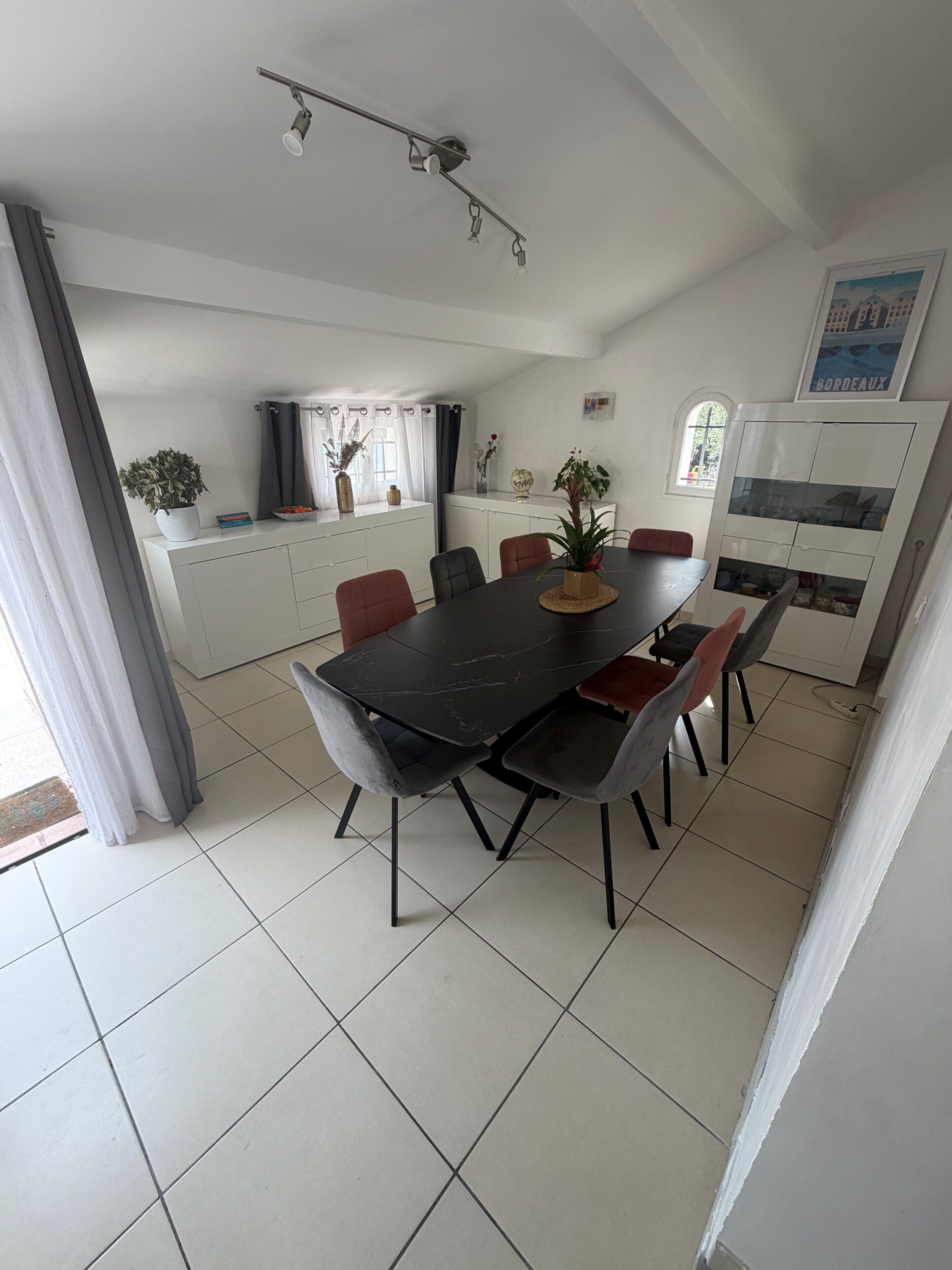 A dining area with a black table and chairs, a ceiling light fixture, and a kitchen area with cabinets and a window.