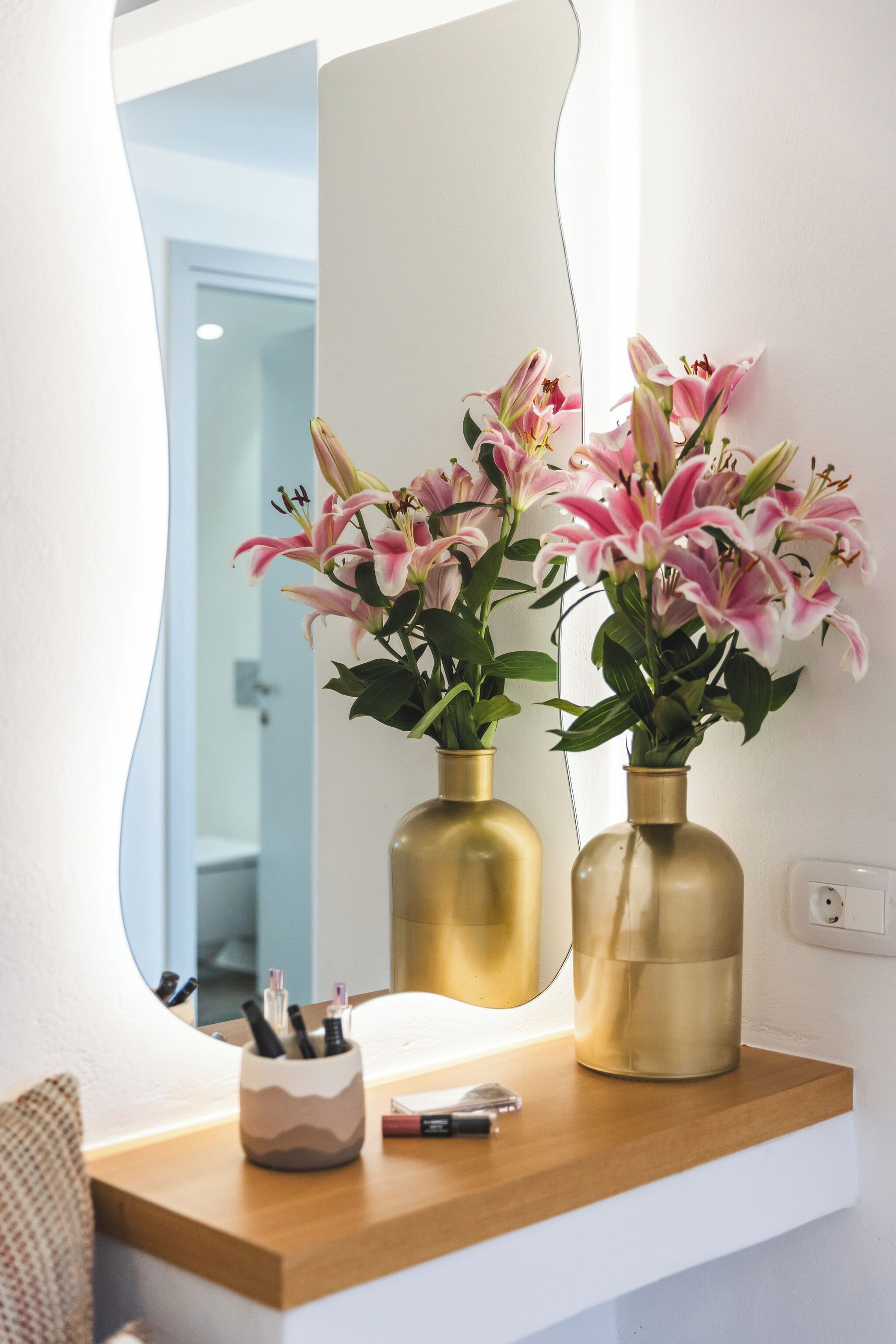 A bathroom vanity with two gold vases of pink lilies, a mirror, and a small container with pens.