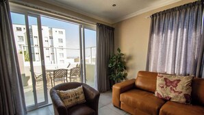 A living room with a brown leather sofa, a brown armchair with a cushion, a potted plant, and a sliding glass door leading to an outdoor area with a table and chairs. - Long Beach Self Catering Studio's (Langstrand)