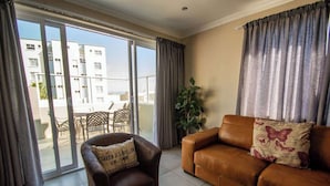 A living room with a brown leather sofa, a brown armchair with a cushion, a potted plant, and a sliding glass door leading to an outdoor area with a table and chairs. - Long Beach Self Catering Two Bedroom Flat (Langstrand)