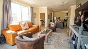 A living room with a sofa, armchair, coffee table, and a dining area with a table and chairs. - Long Beach Self Catering Two Bedroom Flat (Langstrand)