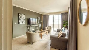 A living room with a sofa, armchair, coffee table, TV, and artwork on the wall. - Long Beach Self Catering Two Bedroom Flat (Langstrand)