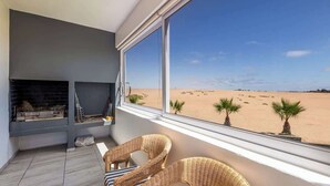 Una habitación con un ventanal grande con vistas a la playa, chimenea, muebles de mimbre y vista a la arena.