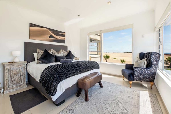 A bedroom with a large bed, a chair, a nightstand, and a view of the beach.