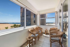 A balcony with wicker furniture, a view of the beach, and a building in the background.
