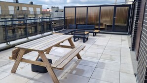 Eine Dachterrasse mit Holztischen und Bänken, einer Glasbrüstung und Blick auf die umliegenden Gebäude.