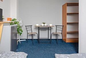 Una habitación pequeña con una cama, un escritorio con dos sillas, un ventilador, una cafetera y una estantería.