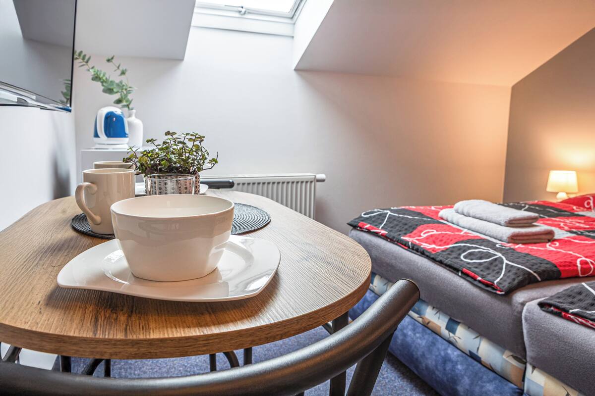 A cozy room with a round wooden table, a bed with a red and black quilt, a potted plant, and a radiator.