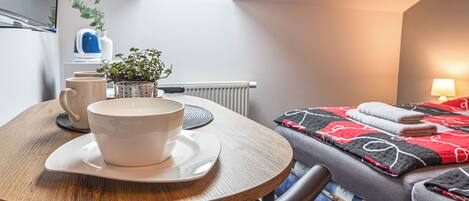 Una habitación acogedora con una mesa redonda de madera, una cama con colcha roja y negra, una planta en maceta y un radiador.