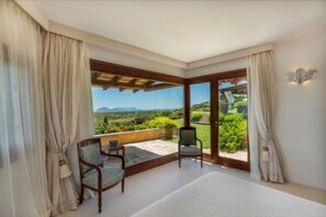 A room with a large sliding glass door leading to a balcony with a view of greenery and mountains. There are two chairs and a small table on the balcony. - ELITE COLLECTION- Cala di Volpe Luxury Villa | Infinity Pool & Sea View (Cala di Volpe)