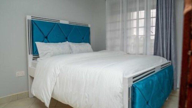 Un lit simple avec un matelas blanc et une tête de lit bleue, dans une pièce aux murs de couleur claire et dotée d’une fenêtre ornée de rideaux légers.