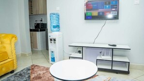 Une pièce moderne avec un fauteuil jaune, une table ronde, un distributeur d’eau, un téléviseur fixé au mur et, au fond, une petite cuisine.